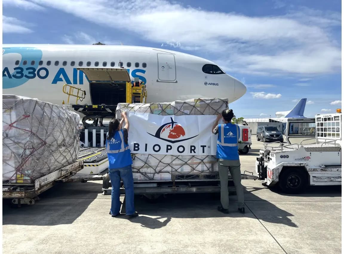 Deux logisticiens de BIOPORT en gilet bleu installant une bâche au logo de l'association sur une grande palette de matériel médical devant un Airbus A330.